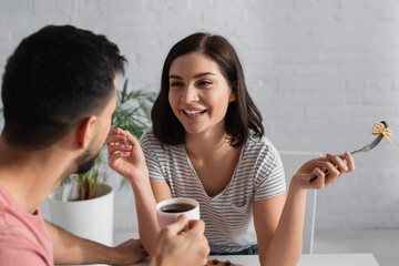 smiling young woman eating pancakes near blurred boyfriend with coffee cup in kitchen