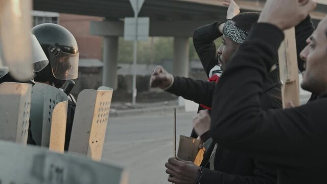 Slowmo tracking shot of riot police officers in full gear holding shields and standing in row while young people chanting and protesting