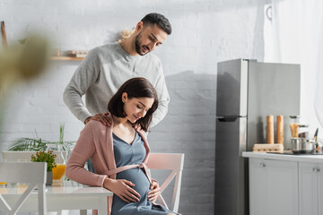 smiling young man holding hands on shoulders of pregnant woman in kitchen