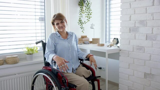 Disabled mature woman in wheelchair packing handmade products indoors at home, looking at camera. Small business concept.