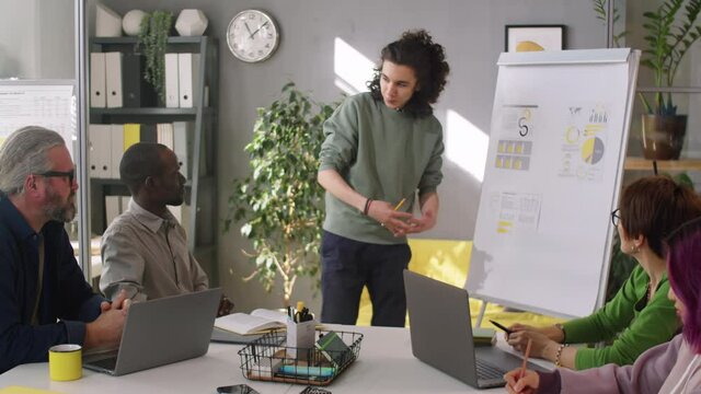 Young male trainee in casualwear standing by flipchart and giving business presentations to multiethnic team of colleagues at office meeting
