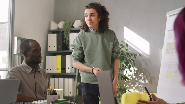 Young man in casualwear standing by flipchart and explaining business presentations to team of diverse colleagues at office meeting