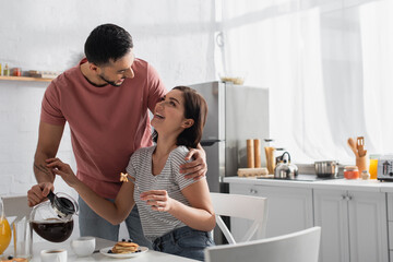 Fototapeta na wymiar smiling young man hugging girlfriend with pieces of pancakes on fork and pouring coffee from pot to cup in kitchen
