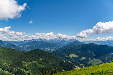 Panoramic view over the Austrian Alps with a hiking trail, trees, and a blue sky with clouds