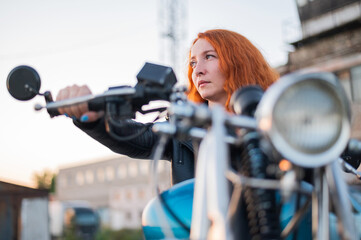 Fototapeta premium Curly red-haired woman in a black leather jacket sits on a motorcycle. Portrait of a serious girl driving a bike.