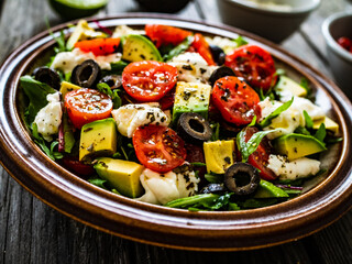 Fresh vegetable salad with mozzarella, tomatoes, avocado, lettuce, black olives and onion on wooden table

