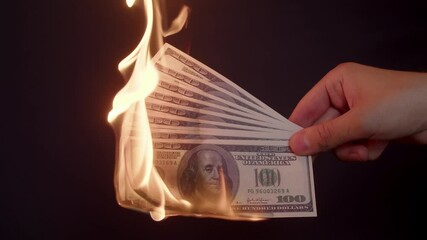 Several hundred-dollar bills in hand, which are slowly burning on a black background in close-up. Hand holds burning dollar bills