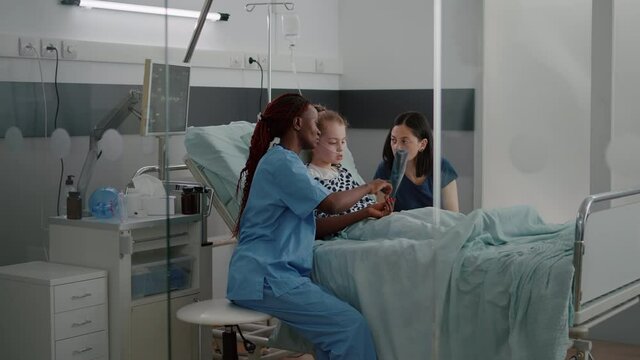 African american nurse showing lungs radiography explaining breathing disease symptoms discussing recovery treatment. Sick little chid resting in bed during clinical examination in hospital ward