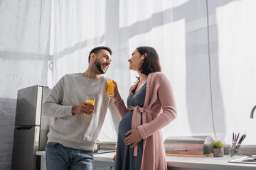 happy young man and pregnant woman standing with orange juice in kitchen