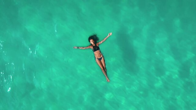 Aerial view an athletic woman lies on her back on blue water. Beautiful slender girl in black swimsuit is resting in warm calm sea, slow motion. Relaxing colorful ocean. Vacation in Panama, San Blas