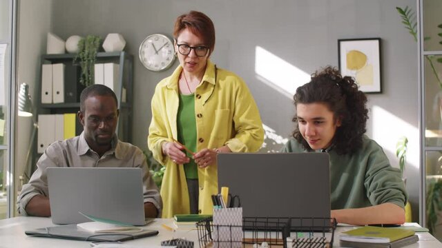 Female business mentor standing in office and giving advice to young multiethnic male trainees as they working on laptops