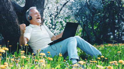 An elderly white-haired man with a laptop is excited, and rejoices in an online victory or good news sitting on the grass among the flowers in the park on a sunny summer day.