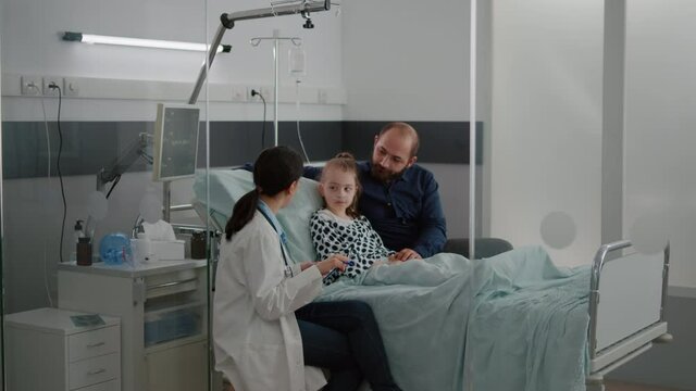 Practitioner woman doctor monitoring sick girl discussing recovery treatment with father in hospital ward during healthcare examination. Child patient resting in bed having breathing disease