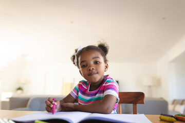 Portrait of happy african american girl sitting at dining table drawing in book, smiling to camera