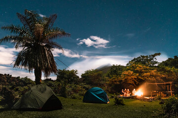 A young group of friends gather around a camp fire at the skirts of a volcano and surrounded by tropical nature.