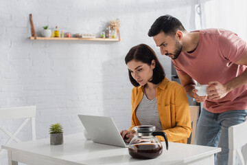 serious young man drinking coffee and looking at laptop near girlfriend in kitchen