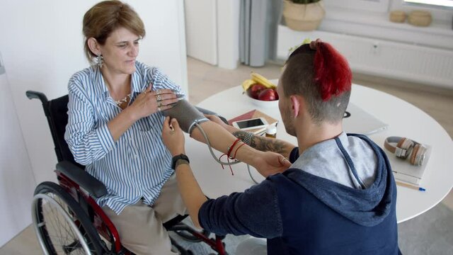 Disabled people in wheelchair sitting at the table indoors at home, measuring blood pressure.