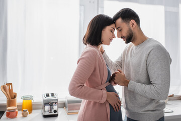 young man with closed eyes gently holding hand of pregnant woman in kitchen