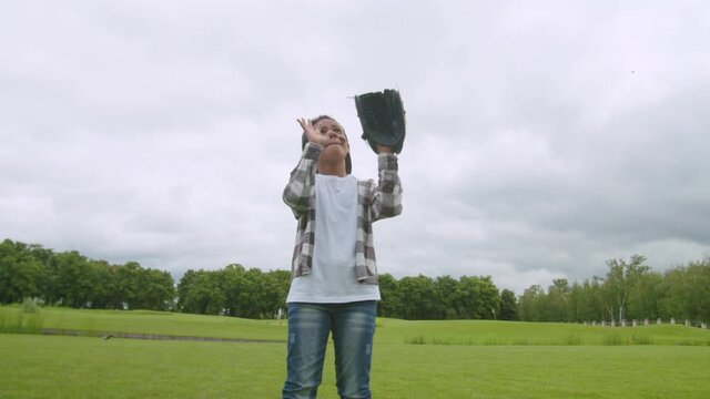 Lovely preadolescent african boy with baseball glove looking up, trying to catch fly ball during baseball game, looking with concentrated expression while playing on green field.