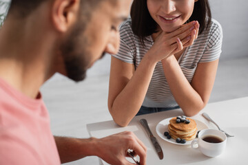 young woman with hands near face seating near blurred boyfriend eating pancakes in kitchen