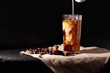Glass cup with cold coffee and ice, coffee beans on a dark background. A hand pours cream.