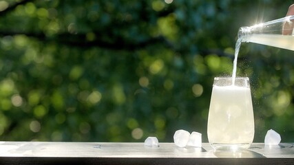 Making drinks outdoors. Hand pours a carbonated drink into a transparent glass with ice. Little splashes fly to the sides from a highly carbonated drink. Many ice cubes in a glass and around - obrazy, fototapety, plakaty