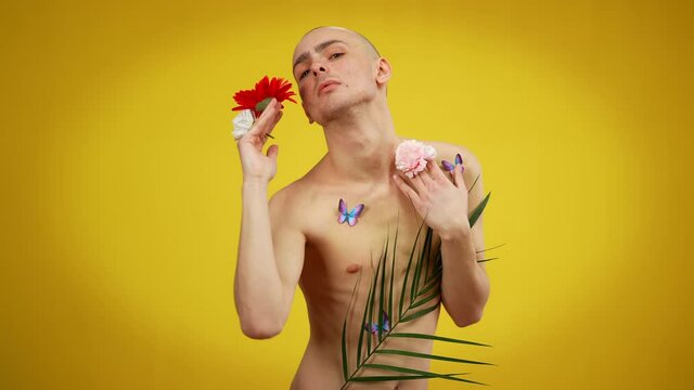 Expressive male queer covered with flowers and butterflies posing at yellow background. Portrait of young Caucasian bisexual man expressing individuality