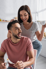 Fototapeta premium smiling young woman touching boyfriend with pieces of pancakes on fork and holding cup with coffee in kitchen