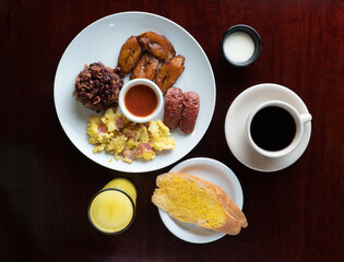 Latin breakfast with fried plantain, scrambled eggs, ground beans, chorizo ​​and orange juice or coffee, all on a white plate