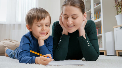Young mother looking at her little son doing homework on carpet in living room. Concept of child education, development and happy parenting.
