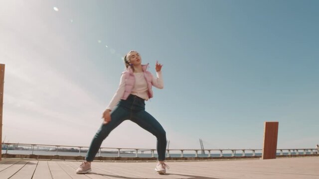 Low angle wide of young Caucasian woman wearing jeans and puffer vest, dancing in modern style on urban deck