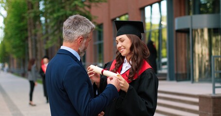 Happy female graduate are embracing with her father with diploma at her hands and rejoicing with each other. Man is hugging his daughter with other students moving and talking in background - obrazy, fototapety, plakaty