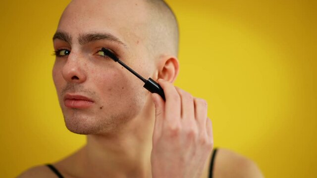 Male Caucasian extravagant transsexual man applying mascara on eyelashes. Close-up portrait of confident male queer doing makeup posing at yellow background. Cosmetics and gender identity