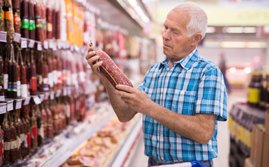 old age man examines salami in sausage department of supermarket