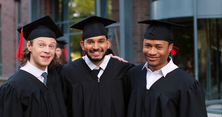 Happy group of mature students on graduation day embracing with each other. Three best friends in academic gowns and caps hugging in front of the camera