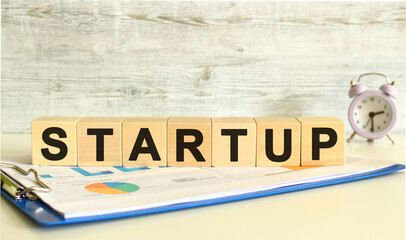 Wooden cubes lie on a folder with financial charts on a gray background. The cubes make up the word STARTUP.