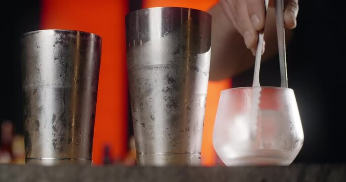 Bartender puts big ice cube to the frozed cocktail glass by forceps, making the cocktail in the bar counter, 4k Prores HQ