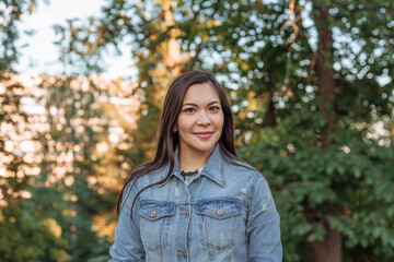 portrait of a beautiful young Asian woman with dark hair in a park.  a pretty Asian woman in a denim jacket against the background of green trees smiles