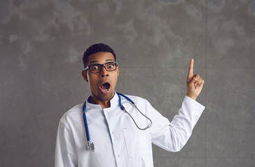 Shocked and concerned african american male doctor in white coat, stethoscope pointing finer up headshot studio portrait on grey background. Healthcare worker, prevent virus, insurance, medicine