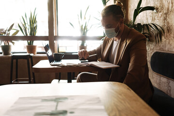 Young white man in face mask working with laptop in cafe