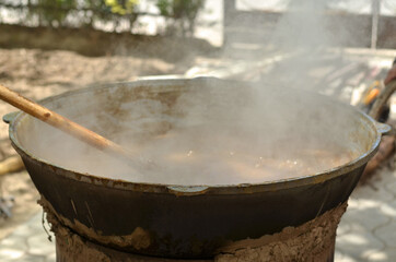 Large black cauldron on the street, a brown food is cooked in it, steam is stirring it with a large wooden spoon, Asian and Uzbek cuisine, cooking food
