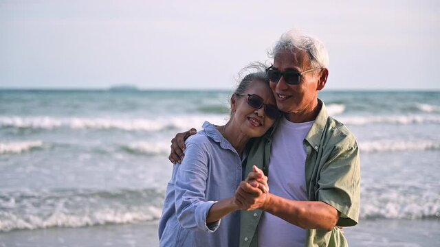 Asian senior couple tourist is hand in hands, embrace and dancing on tropical sea beach in summer holiday. Happy family couple elderly older retire resting relax together lifestyle on seaside outdoor.