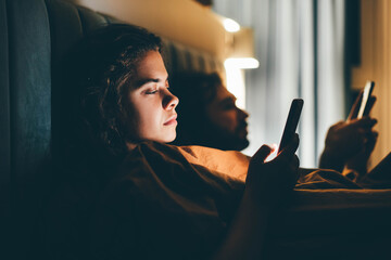 Couple with smartphones in their bed. Mobile phone addiction. Bored distant couple ignoring each other lying in bed at night while using mobile phones. - obrazy, fototapety, plakaty