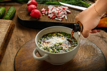 A woman's hand pours kvass from a bottle into okroshka in a tureen with herbs and an egg.