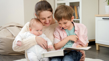 Smiling woman hugging her baby and older son while reading story book at living room. Parenting, children happiness and family relationship