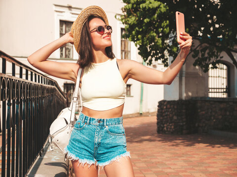 Portrait of young beautiful smiling hipster woman in trendy summer jeans shorts and hat. Sexy model posing on the street background at sunset. Positive model outdoors with briefcase.Taking selfie