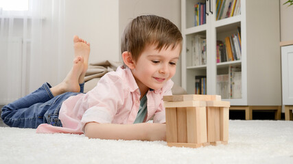 Cute smiling boy lying on floor and looking at toy house he built from toy wooden blocks. Concept of child education and games at home