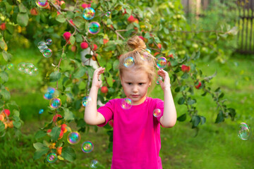 Portrait of children in apple orchard. Little girl in pink Tshirt stands near branches of an apple tree, covering her face with her hands, hiding from soap bubbles. Carefree childhood, happy child