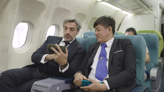 Two senior Asian and caucasian businessman using smartphone consulting and talking about business plan and result during traveling abroad by business class plane. Transportation or business concept.