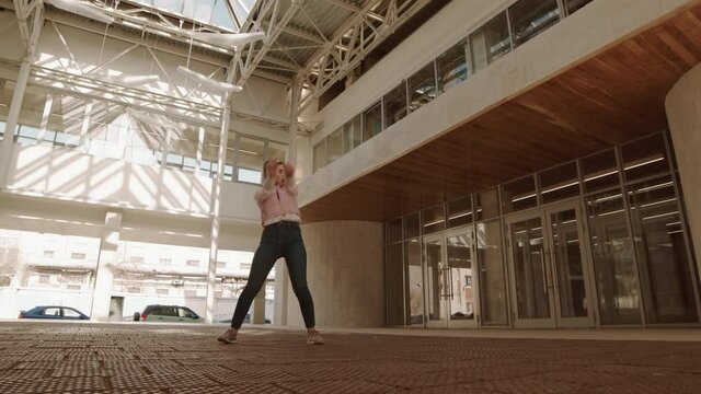Wide shot of joyful Caucasian young woman wearing casual jeans and puffer jacket, dancing in premises of abandoned semi-outdoor mall